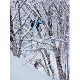 Burton Custom Snowboard 2025 - A snowboarder is riding a blue and white snowboard with black lettering, jumping in front of bare trees covered in snow.