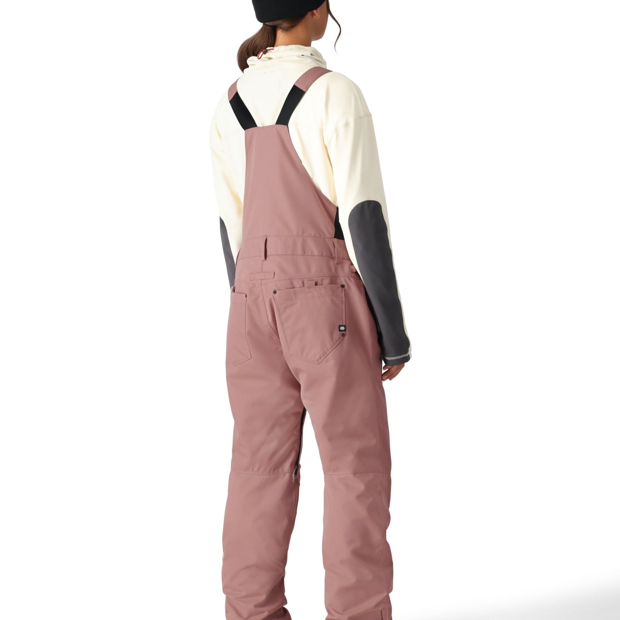 The woman in the black beanie and pink bib overalls has her back to the camera, wearing a cream-colored turtleneck sweater underneath.