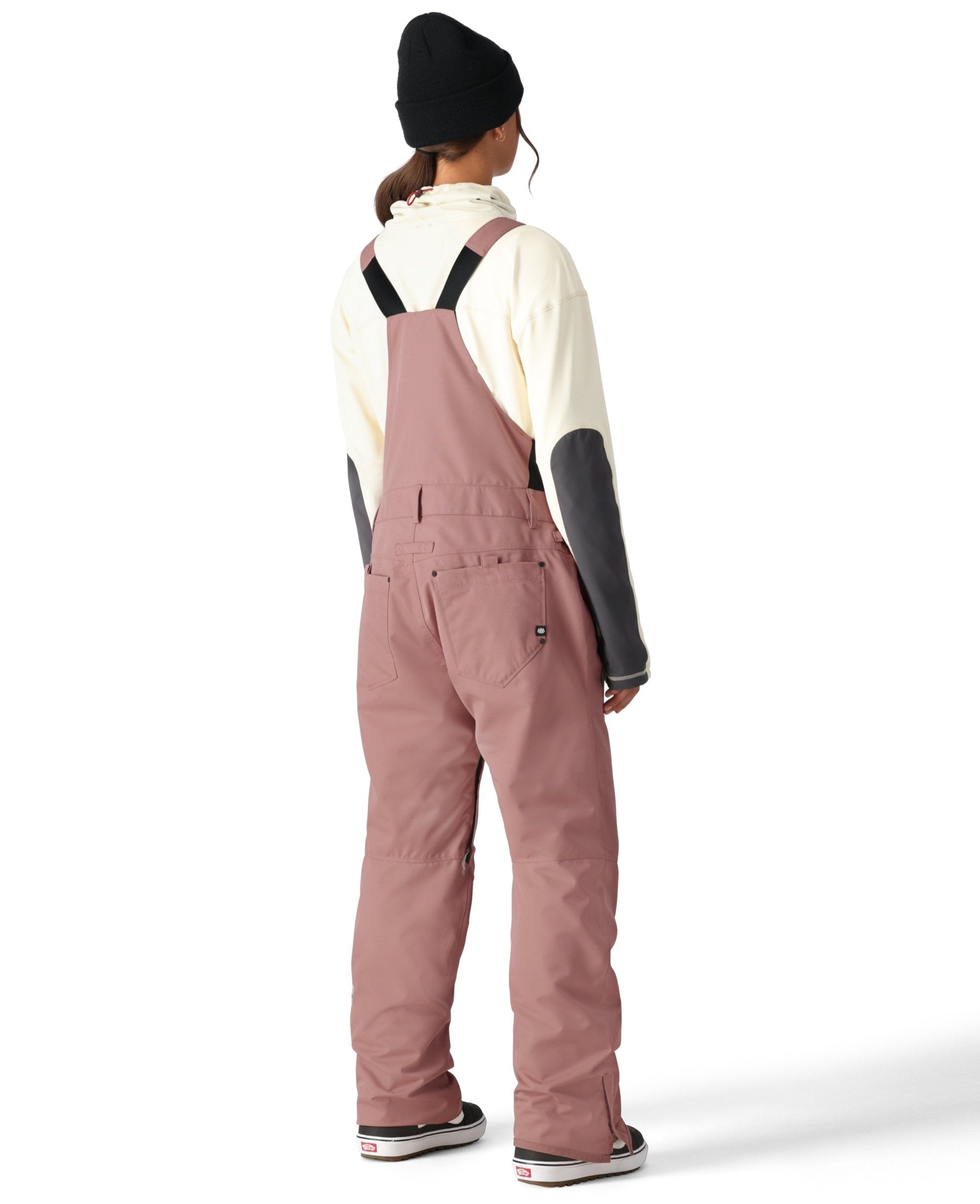 The woman in the black beanie and pink bib overalls has her back to the camera, wearing a cream-colored turtleneck sweater underneath.