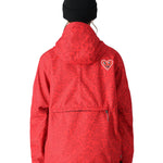 The 686 Women's Outline Shell Anorak 2025 is showcased in this image, wearing a vibrant red jacket with black accents and a heart-shaped logo on the left sleeve.