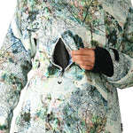 The 686 Whisper Insulated Jacket for women has a white base with a black leaf pattern and is being zipped up by a woman.