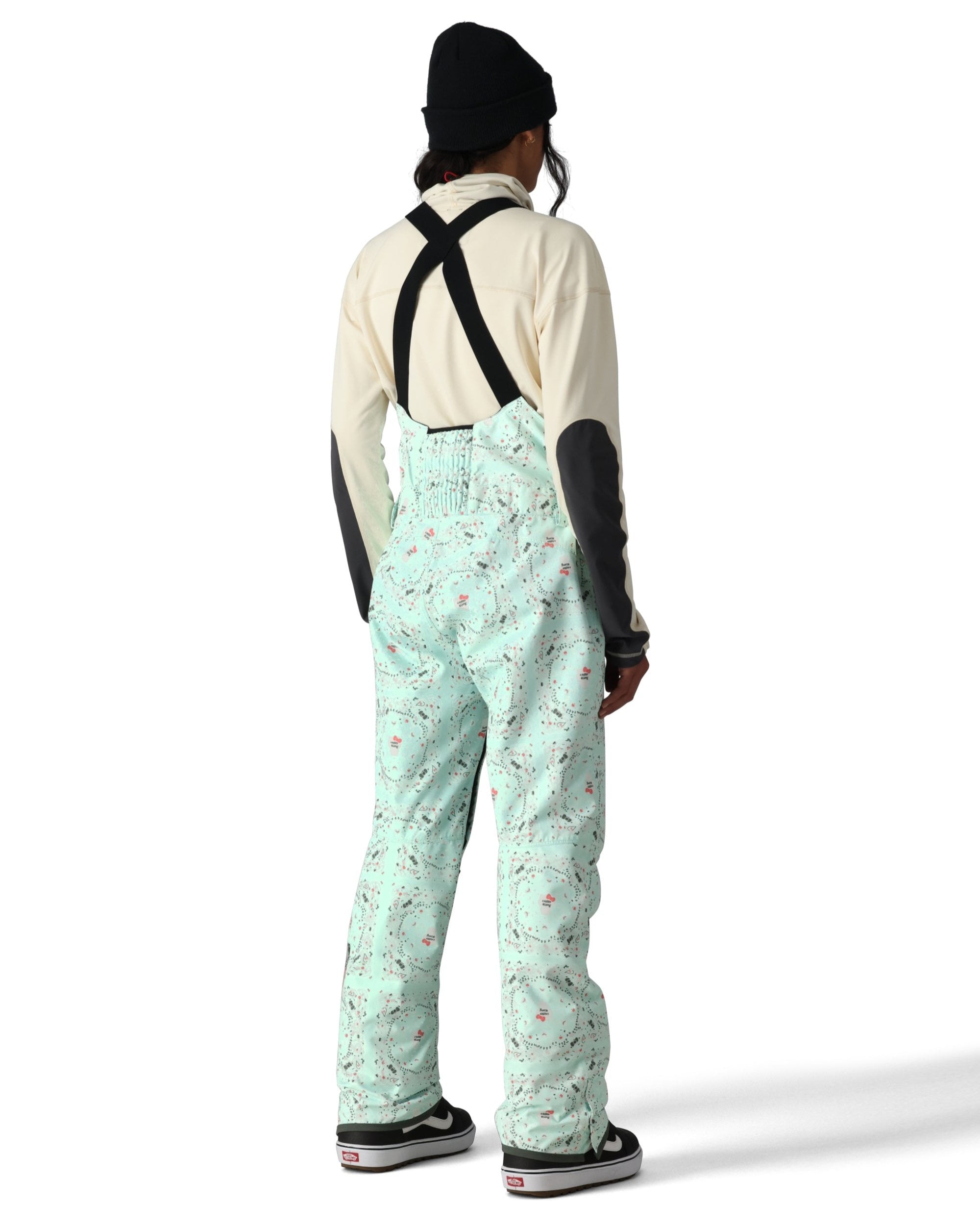 The 686 Harper Shell Bib pants are light green with a pattern of gray and pink ski trails on them.