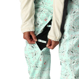 The 686 Harper bib pants have a light blue-green color with a pattern of small pink and gray shapes, and are being modeled in the front view.