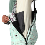 The 686 Women's Harper Shell Bib 2025 is primarily mint green with a pattern of small black and red shapes on its front view.