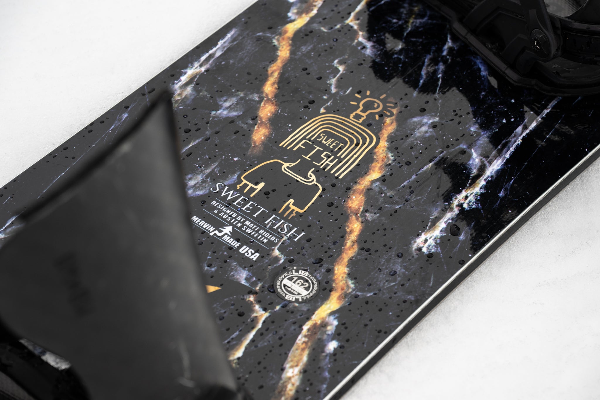 Lib Tech Mayhem Sweetfish snowboard deck close-up, gold tribal logo on black marbled base