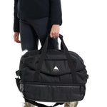 Burton Gig 70L Duffel Bag - A large black duffel bag with a white logo featuring a mountain peak and the word "BURTON" in small letters, held by a person wearing dark pants.