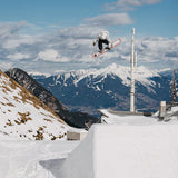 Burton Good Company B Grade Snowboard 2024 - A snowboarder in mid-air, wearing a black and white jacket, with a snowy mountain range in the background.