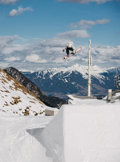 Burton Good Company B Grade Snowboard 2024 - A snowboarder in mid-air, wearing a black and white jacket, with a snowy mountain range in the background.