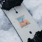 Burton Good Company Snowboard 2025 - A white snowboard with a gold border around a giraffe standing in front of a red desert landscape.