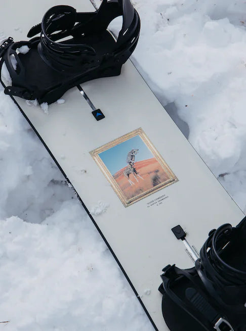 Burton Good Company Snowboard 2025 - A white snowboard with a gold border around a giraffe standing in front of a red desert landscape.
