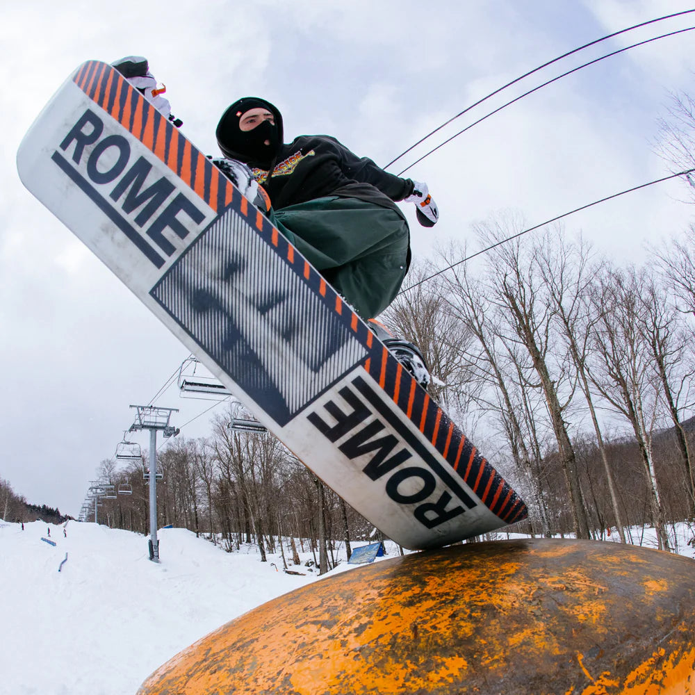 Rome Agent Snowboard 2025 - A snowboarder is riding a snowbank, with their board facing left and slightly upwards. The board has an orange stripe along its top edge.