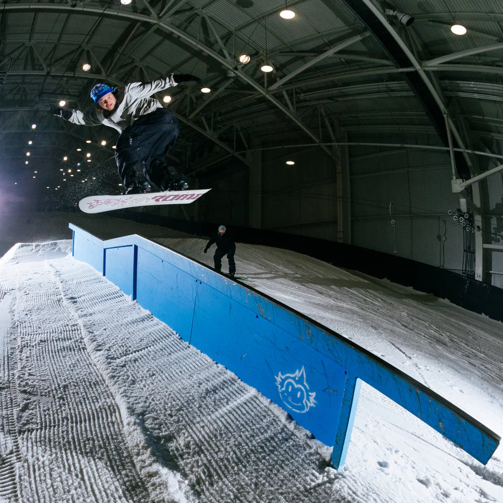 Rome Rene-Gade Snowboard 2025 - A snowboarder in mid-air, wearing a white jacket and black pants, with a blue helmet, riding over a blue rail on a snowy slope.