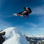 Rome Stale Crewzer Snowboard 2025 - A snowboard with a white and orange lightning bolt graphic on its bottom, being ridden by a person in mid-air above a snowy mountain landscape.