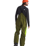 North Face Men's Ceptor Shell Bib - A man wearing a pair of olive green bib overalls with neon yellow accents on the side seams and black boots.