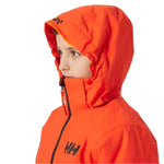 Helly Hansen Junior's Traverse Insulated Jacket - A young boy wearing a bright orange jacket with black lettering and a zipper, looking to the left side of the image.