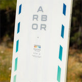 Arbor Women's Mantra Rocker Snowboard 2024 - A white snowboard with blue and green geometric shapes on the top and bottom edges.