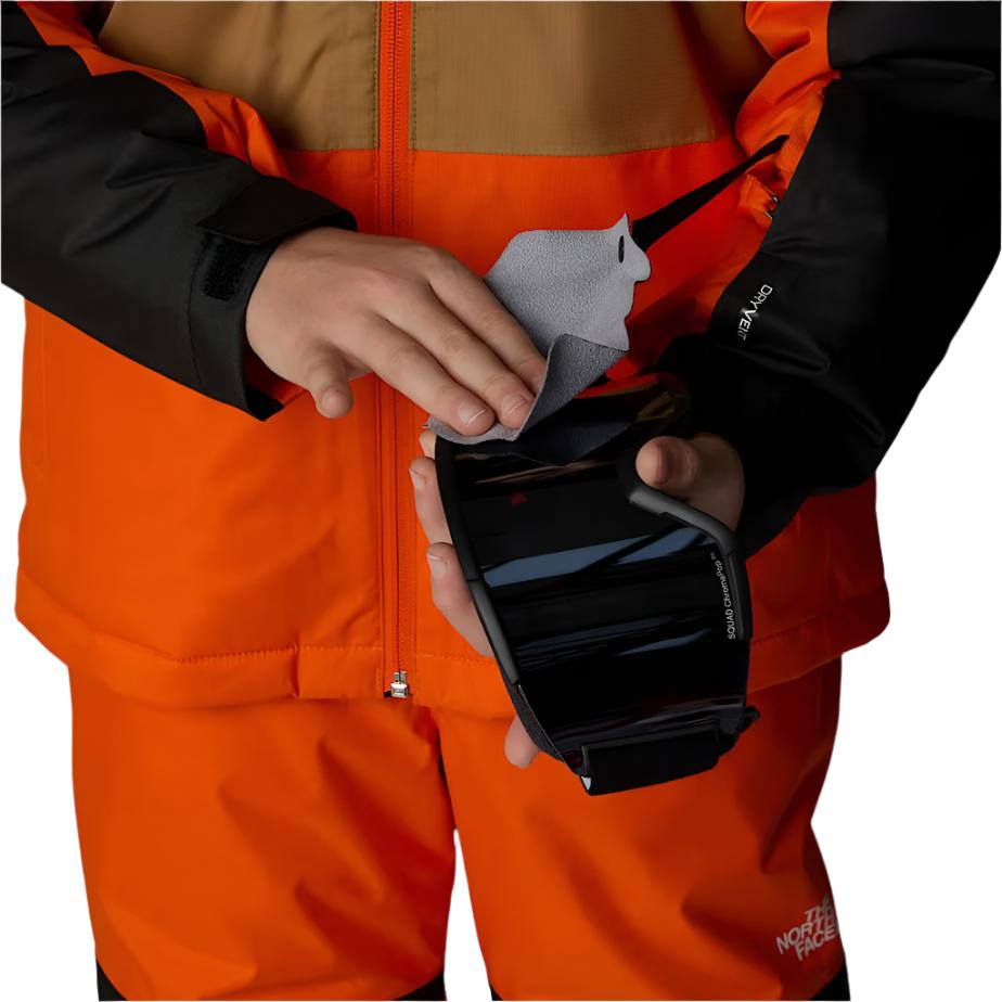 North Face Boys' Freedom Insulated Jacket - A boy wearing a bright orange and tan jacket with black sleeves, holding a gray fleece item in his hand.