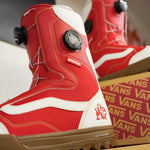 Vans Women's Encore Pro Limited Edition Snowboard Boots 2026 - A pair of red snowboard boots with white accents and a black BOA dial on top, sitting in front of a cardboard box with the word "VANS" printed on it.