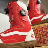 Vans Women's Encore Pro Limited Edition Snowboard Boots 2026 - A pair of red snowboard boots with white accents and a black BOA dial on top, sitting in front of a cardboard box with the word "VANS" printed on it.
