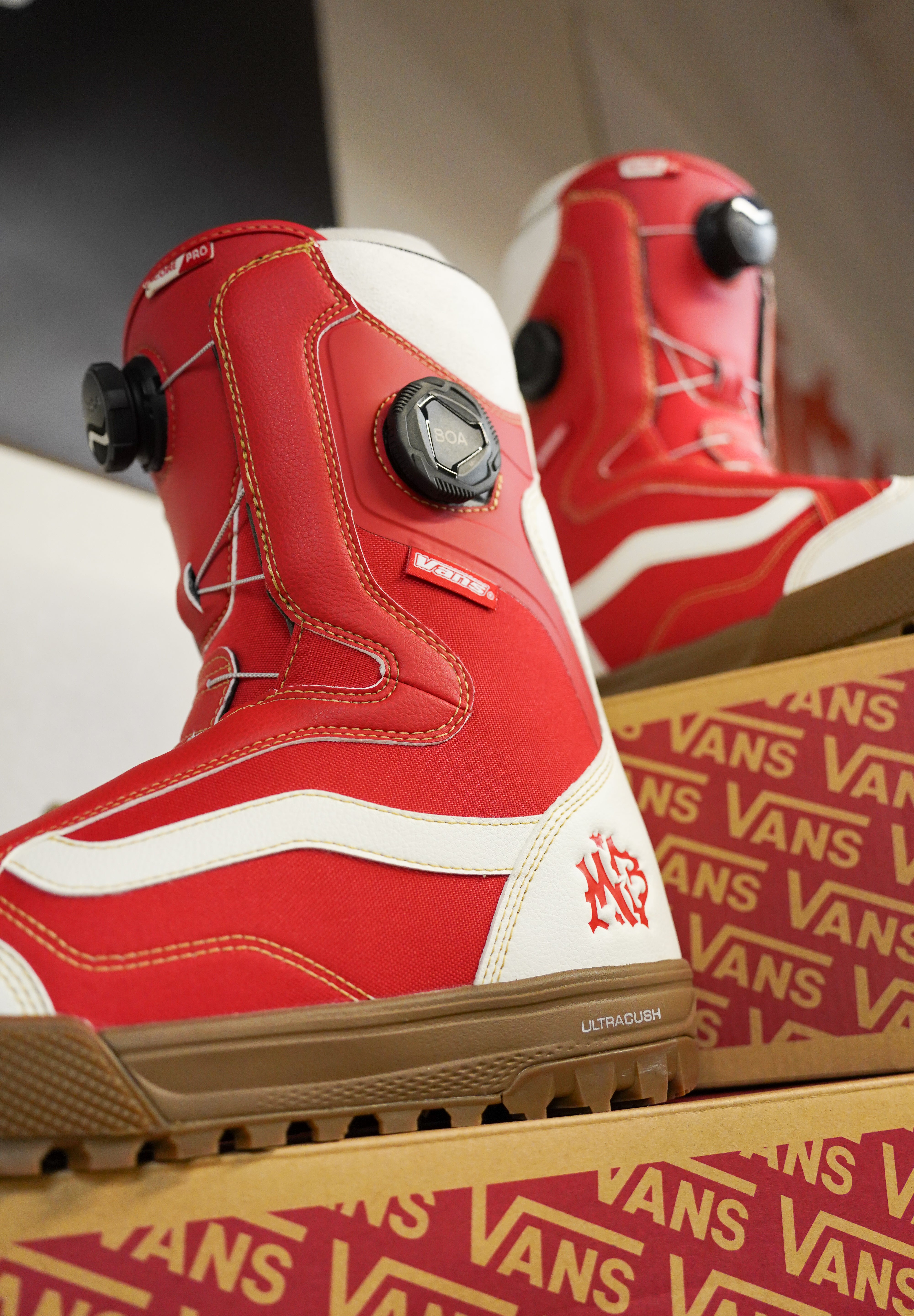 Vans Women's Encore Pro Limited Edition Snowboard Boots 2026 - A pair of red snowboard boots with white accents and a black BOA dial on top, sitting in front of a cardboard box with the word "VANS" printed on it.