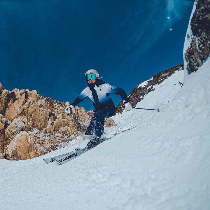 Descente Women's Swiss Insulated Jacket - A woman in a white and blue jacket is skiing down a snowy mountain, wearing a helmet with goggles.