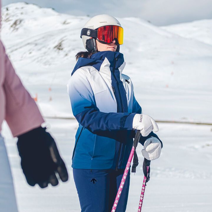Descente Women's Swiss Insulated Jacket - A woman in a white and blue jacket with a hood, wearing a white helmet and red goggles, holding ski poles while standing on a snowy mountain.
