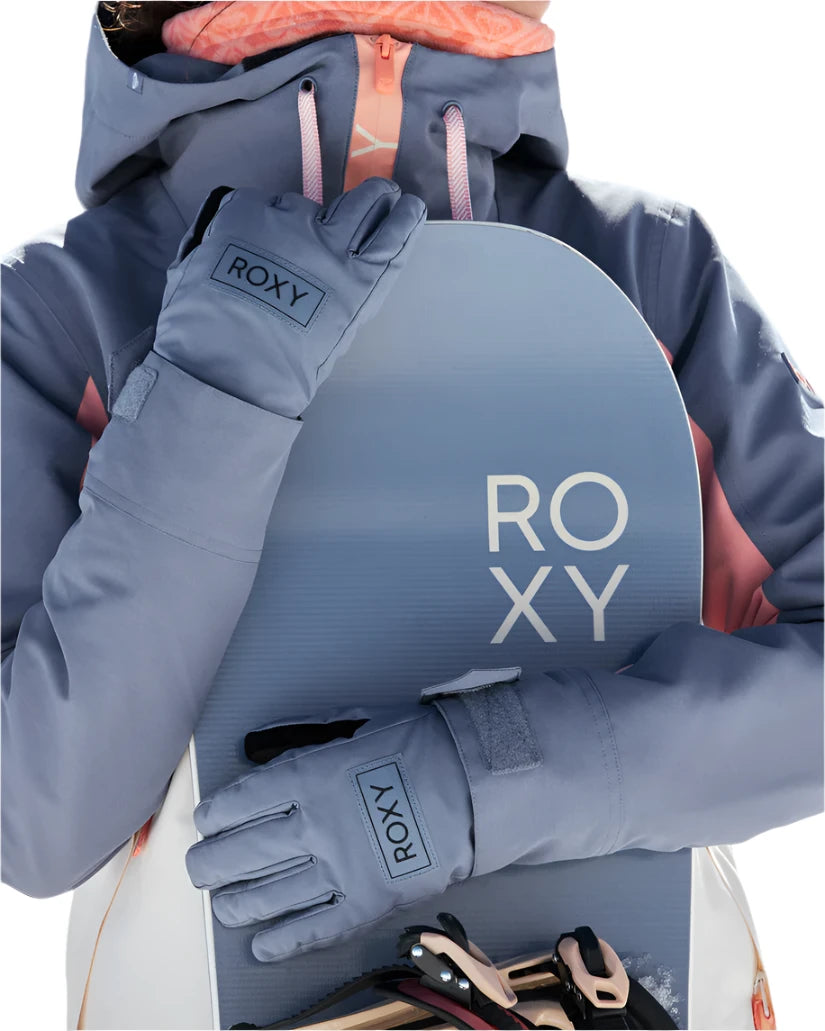 Roxy Women's Freshfield Glove 2025 - A woman wearing a gray snowboard jacket with pink accents holds a light blue snowboard in front of her torso, showcasing the glove on her left hand.