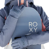 Roxy Women's Freshfield Glove 2025 - A woman wearing a gray snowboard jacket with pink accents holds a light blue snowboard in front of her torso, showcasing the glove on her left hand.
