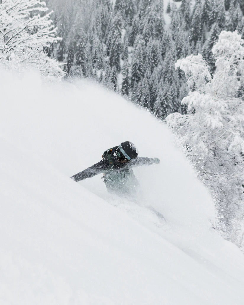 Roxy Women's Stated Insulated Jacket 2025 - A woman wearing a black jacket with brown accents and a backpack, standing in front of snow-covered trees.