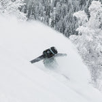 Roxy Women's Stated Insulated Jacket 2025 - A woman wearing a black jacket with brown accents and a backpack, standing in front of snow-covered trees.