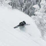 Roxy Women's Stated Insulated Jacket 2025 - A woman wearing a black jacket with brown accents and a backpack, standing in front of snow-covered trees.