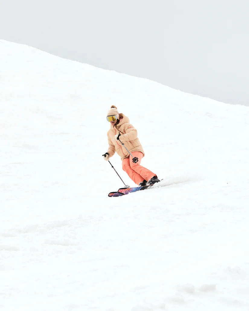Roxy Women's Alofted Puffy Insulated Jacket - A woman in a tan puffer jacket with brown fur trim, wearing goggles and snow pants, skiing down a snowy hill.