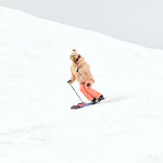 Roxy Women's Alofted Puffy Insulated Jacket - A woman in a tan puffer jacket with brown fur trim, wearing goggles and snow pants, skiing down a snowy hill.