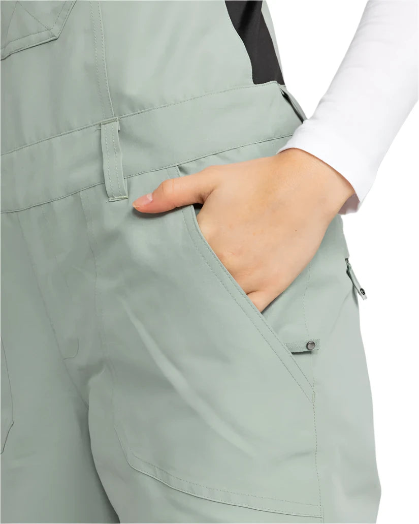 Roxy Women's Rideout Insulated Bib Pant 2025 - A pair of light gray snow pants with a black stripe down the side, featuring a zippered pocket on the thigh and a snap button closure at the waist.