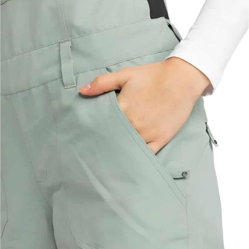 Roxy Women's Rideout Insulated Bib Pant 2025 - A pair of light gray snow pants with a black stripe down the side, featuring a zippered pocket on the thigh and a snap button closure at the waist.