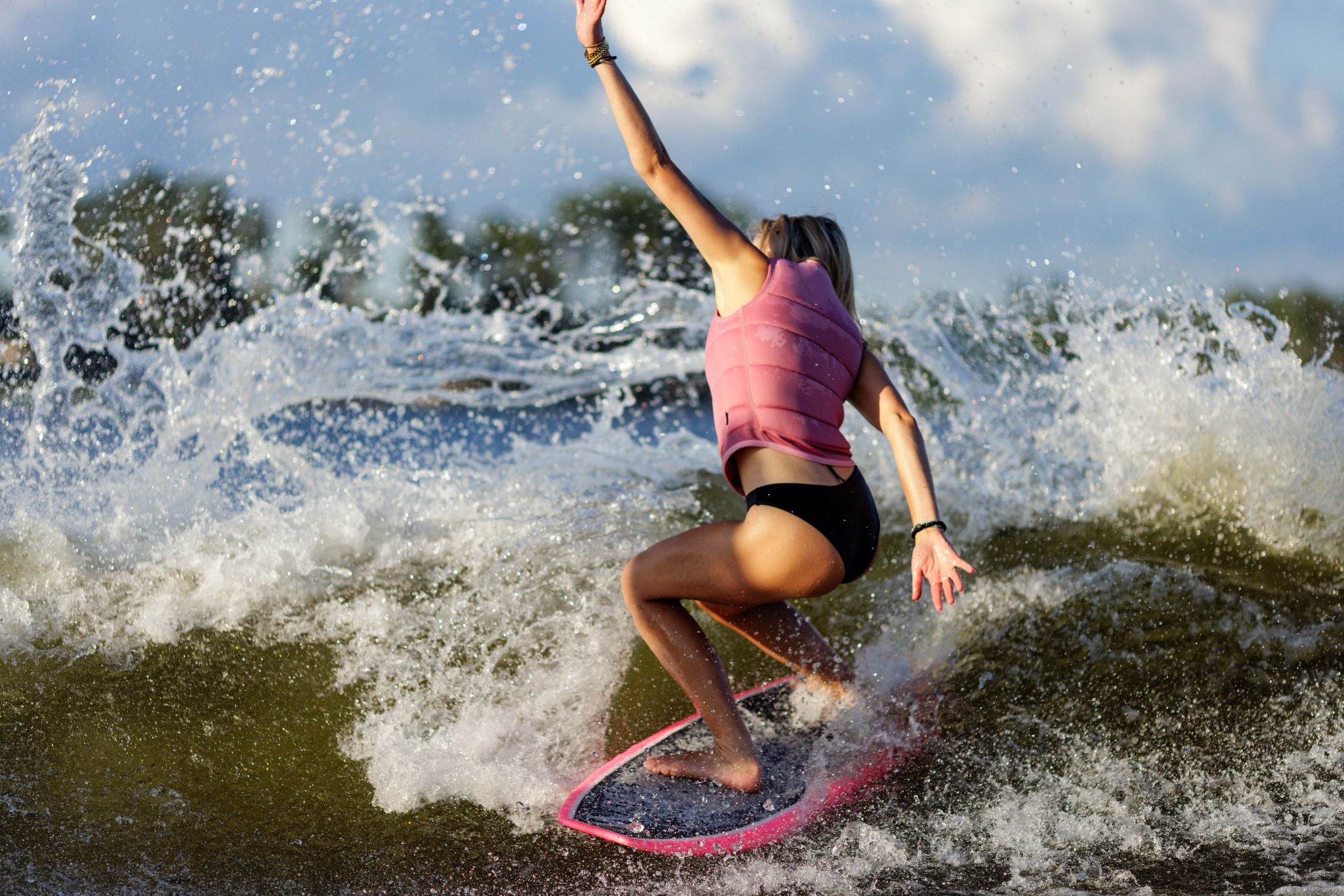 Follow Women's Corp Comp Vest 2024 - Pink vest with black straps on a woman in motion, riding a wave with her arms raised and left leg bent.
