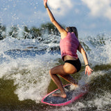 Follow Women's Corp Comp Vest 2024 - Pink vest with black straps on a woman in motion, riding a wave with her arms raised and left leg bent.