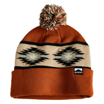 Autumn Surplus Blanket Pom Beanie 2025 - A rust-colored beanie with a large, multicolored pom-pom on top and a cream-colored band featuring a black geometric pattern.