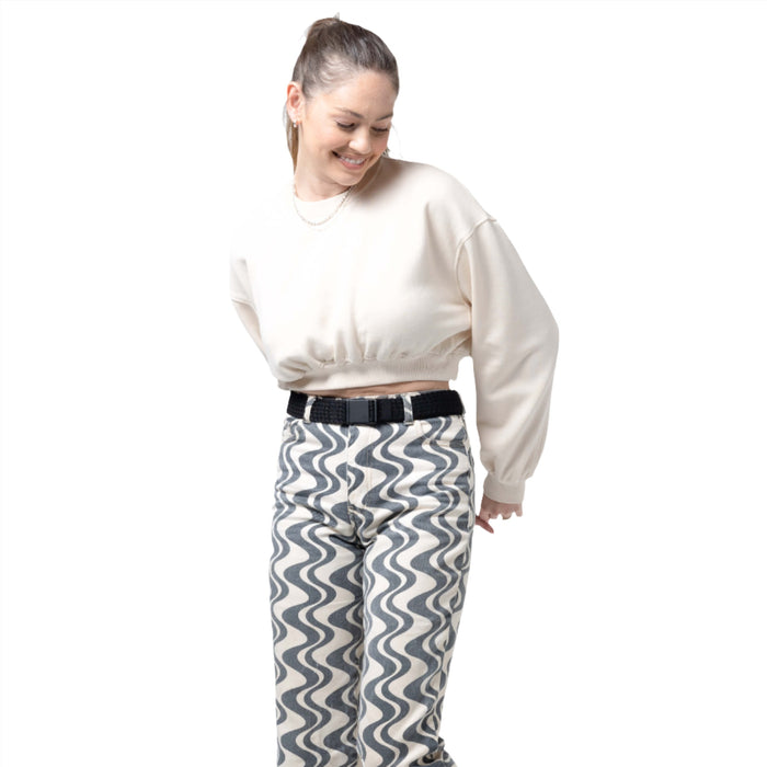 A woman wearing a cream-colored cropped sweater and high-waisted pants with a black and white wavy pattern. She is wearing a black woven belt with a black buckle and smiling while looking down.