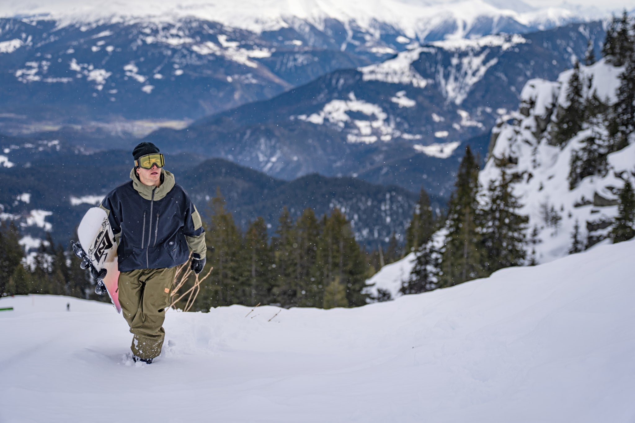 Volcom V.Co WFO Shell Jacket - A man in a black beanie, yellow goggles, and olive green snow pants is walking up a snowy hill while holding a white snowboard with black lettering. He's wearing a navy blue jacket with an olive green hood and olive green accents on the sleeves.