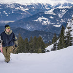 Volcom V.Co WFO Shell Jacket - A man in a black beanie, yellow goggles, and olive green snow pants is walking up a snowy hill while holding a white snowboard with black lettering. He's wearing a navy blue jacket with an olive green hood and olive green accents on the sleeves.