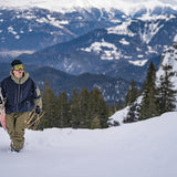 Volcom V.Co WFO Shell Jacket - A man in a black beanie, yellow goggles, and olive green snow pants is walking up a snowy hill while holding a white snowboard with black lettering. He's wearing a navy blue jacket with an olive green hood and olive green accents on the sleeves.