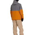 Volcom Men's Rain GORE-TEX Bib Overall - A man wearing a grey and orange rain jacket, khaki bib overalls with cargo pockets, black gloves, and black sneakers.