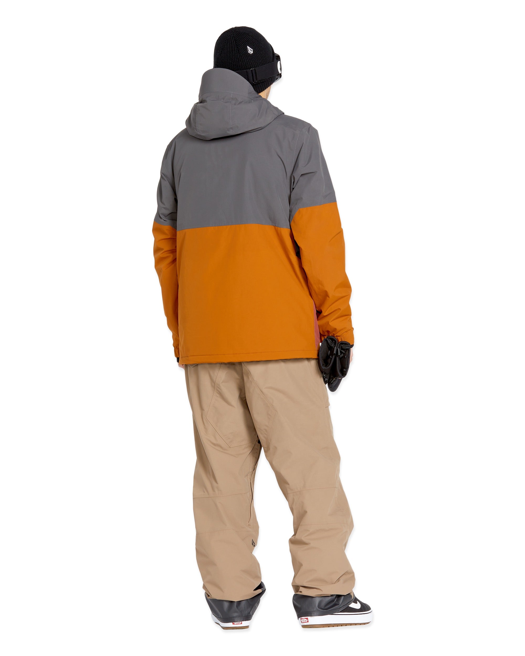 Volcom Men's Rain GORE-TEX Bib Overall - A man wearing a grey and orange rain jacket, khaki bib overalls with cargo pockets, black gloves, and black sneakers.