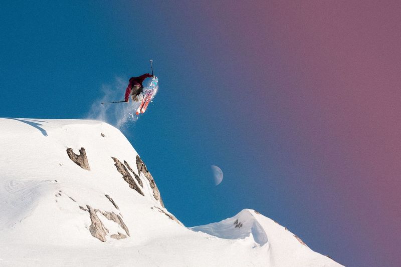 A skier wearing a red jacket is seen skiing downhill on a mountain. The skis are Salomon S/Lab QST Blank Skis 2026, and the person is in mid-air while performing a jump.