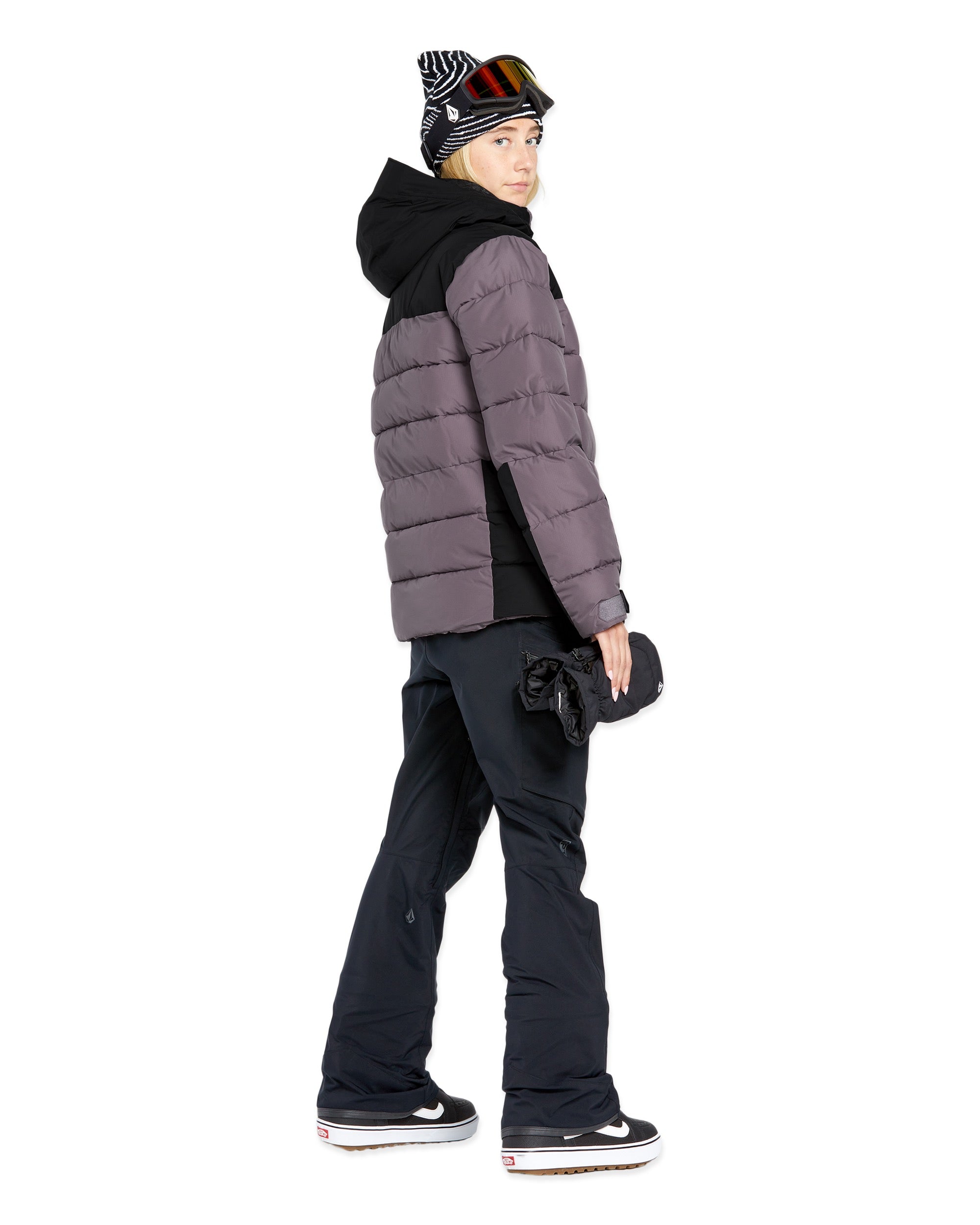 Volcom Women's Elm Stretch GORE-TEX Bib Overall - A woman wearing a black and white striped beanie, a gray puffer jacket with a black hood, and black snow pants with a zippered pocket on the thigh.