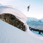 Nidecker Alpha Snowboard 2025 - A white snowboard with a black stripe down the middle, being ridden by a person in mid-air above a snowy mountain slope.
