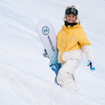 Nidecker Women's Astral Snowboard 2025 - A white snowboard with blue stripes and a black logo on top, held by a woman in a yellow jacket.