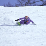 Nidecker Blade Snowboard 2025 - A snowboarder in a purple snowsuit, wearing an orange helmet and goggles, is riding down a snowy slope on a green snowboard with black bindings.