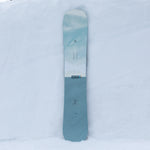 Nidecker Escape Snowboard 2025 - A light blue snowboard with a white stripe and forest green trees printed on it.